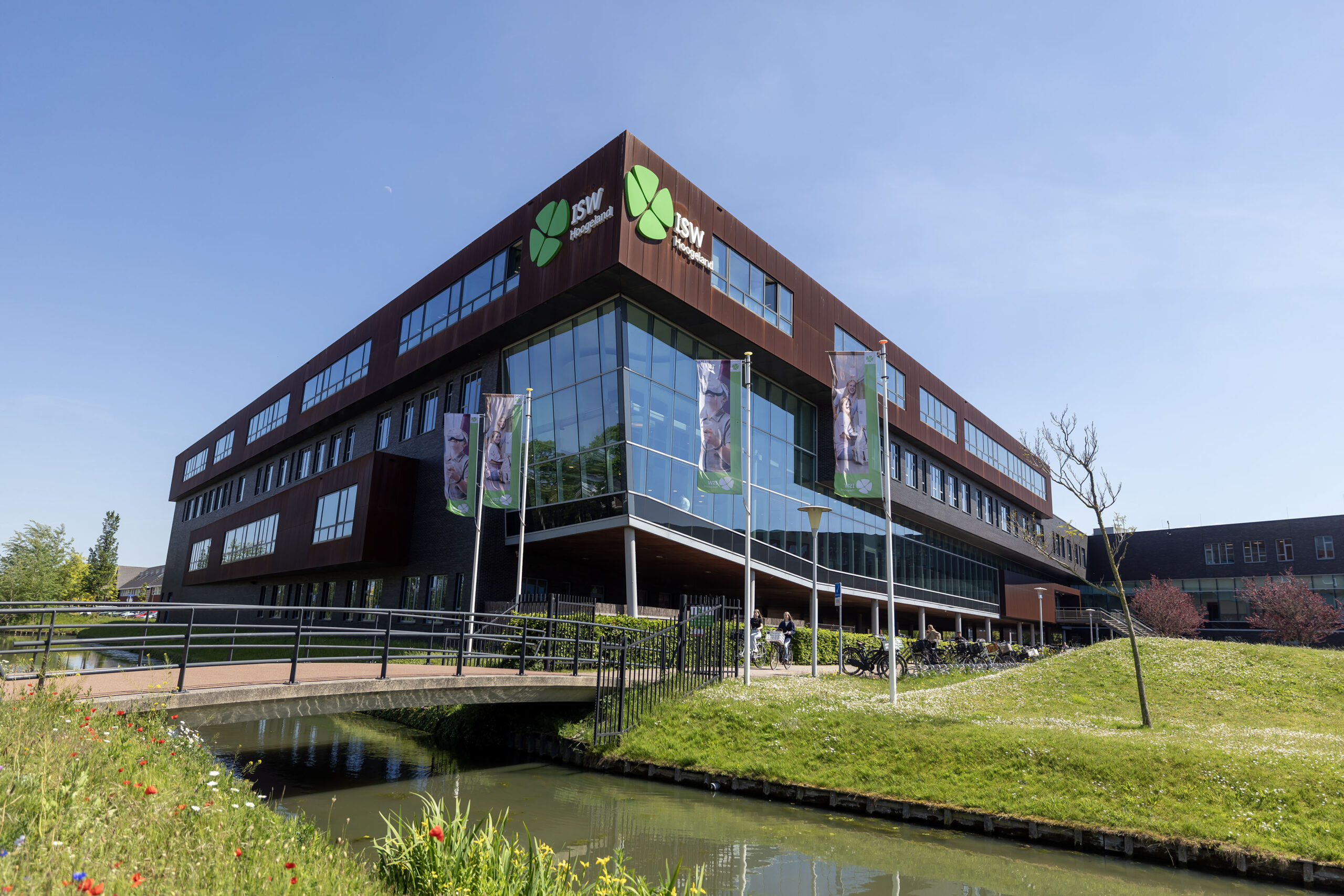ISW - Modern kantoorgebouw met grote glazen ramen en groene banieren, omgeven door gras, een kanaal en een blauwe lucht. Op de voorgrond steekt een loopbrug het kanaal over bij ISW Hoogeland.