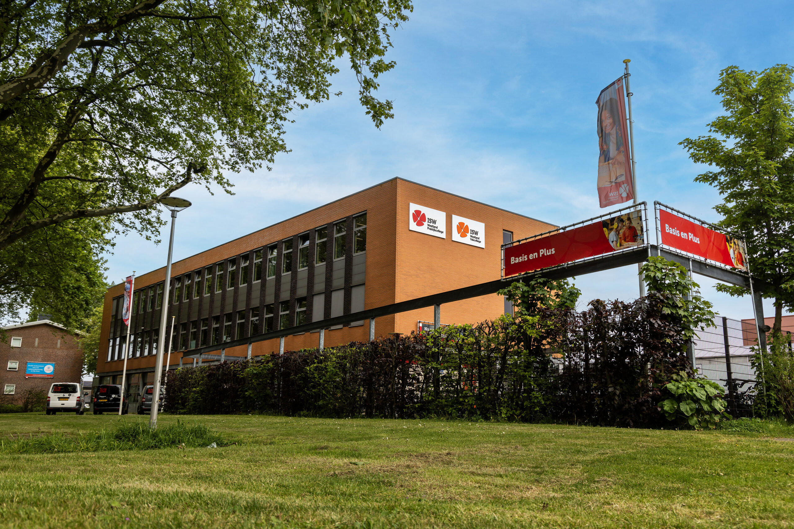 ISW - Een modern bakstenen gebouw met grote ramen en rood-witte borden van het Westland Praktijkcollege bij het dak, omringd door groene bomen en een grasveld onder een strakblauwe hemel.