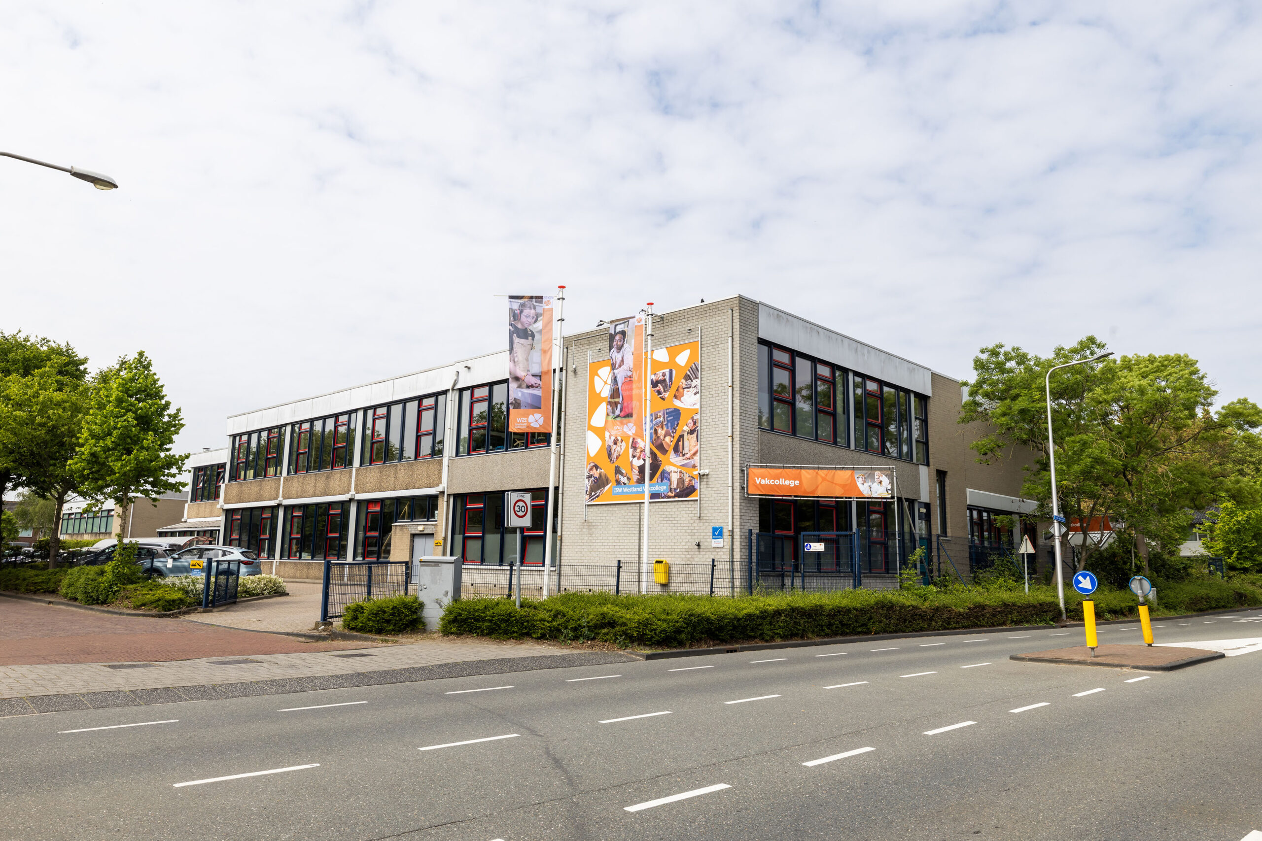 ISW - Een modern gebouw van twee verdiepingen met grote ramen, spandoeken en een oranje bord met Voedselbank op de gevel; het gebouw ligt aan een weg met bomen en een trottoir, vlakbij het Westland Vakcollege.