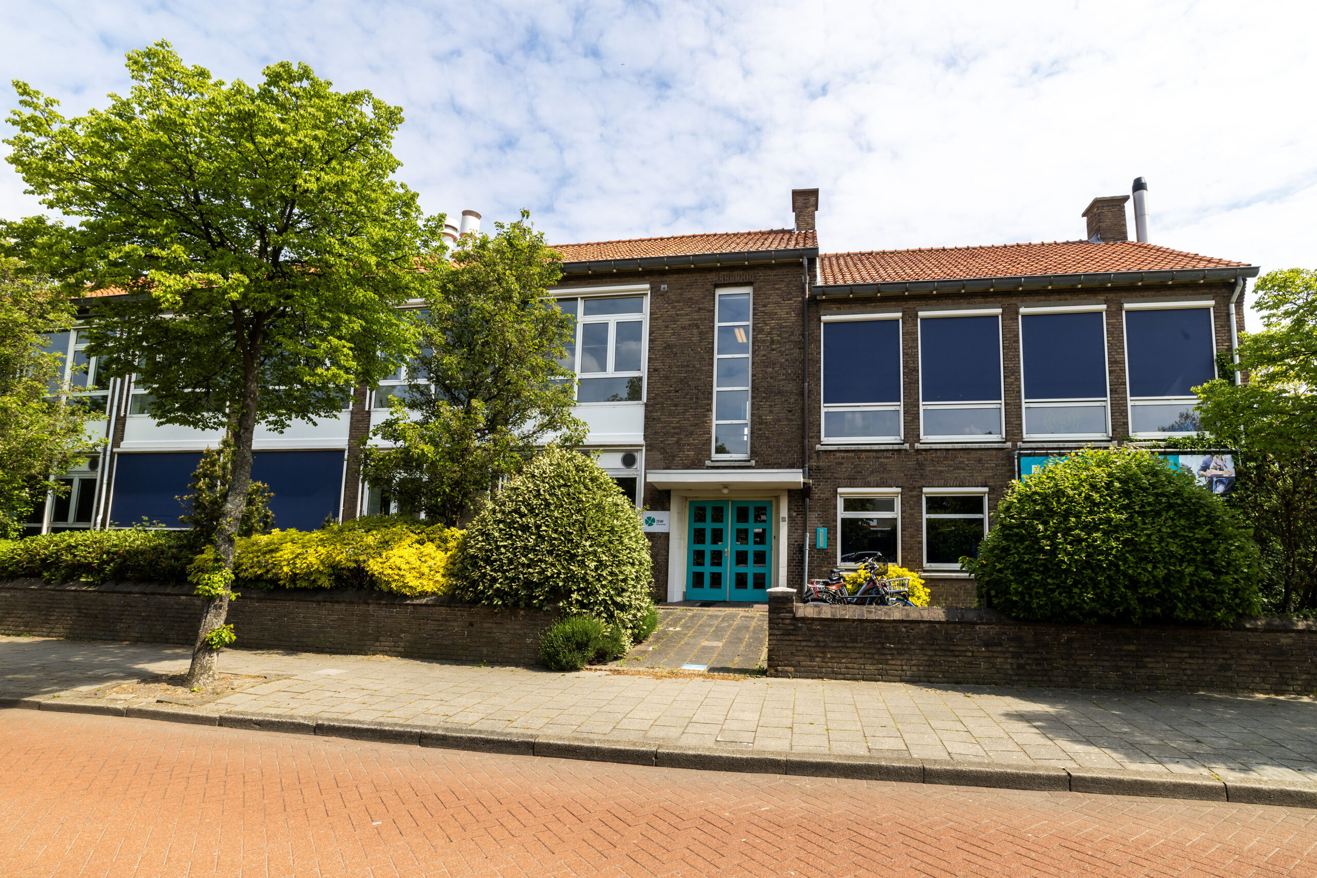 ISW - Een modern bakstenen gebouw met grote blauwgetinte ramen en een turkooizen deur staat aan de ISW Irenestraat, omringd door groene bomen en struiken, met aan de voorkant een trottoir en een straat van rode baksteen.