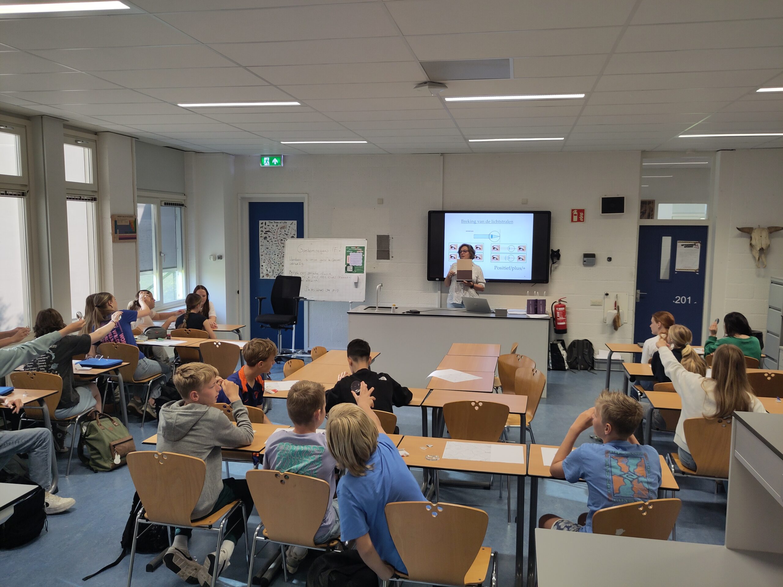 ISW - Een docent staat vooraan in een lokaal van een ISW-locatie en geeft les aan leerlingen die aan hun bureau zitten. Verschillende leerlingen steken hun handen op terwijl er een presentatie op een scherm achter de docent wordt getoond.