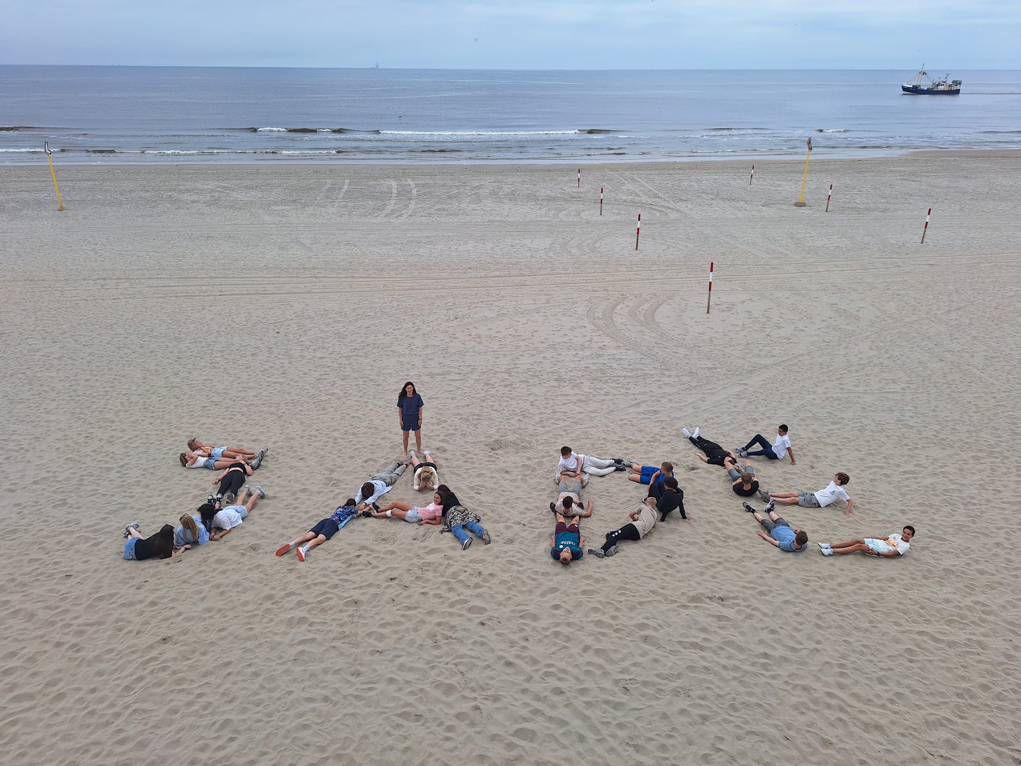 ISW - Een groep leerlingen van ISW Irenestraat en Stichting Jade liggen op een zandstrand en vormen met hun lichamen het woord JAZZ. Eén persoon staat in het midden van de A. De zee en een schip verschijnen onder een bewolkte hemel op de achtergrond.