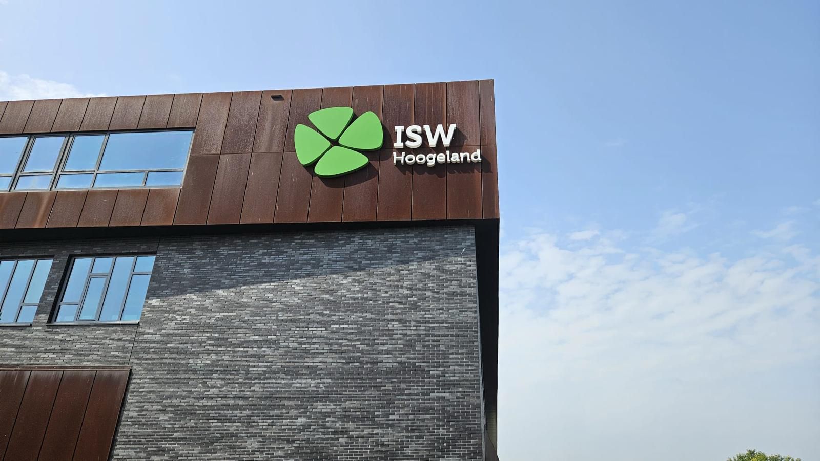 ISW - Een modern gebouw met donkere bakstenen en bruine panelen, onderdeel van de ISW scholen, heeft een groen klaverachtig logo en de tekst ISW Hoogeland op de bovenste gevel, wat het een nieuwe uitstraling geeft tegen de helderblauwe lucht.