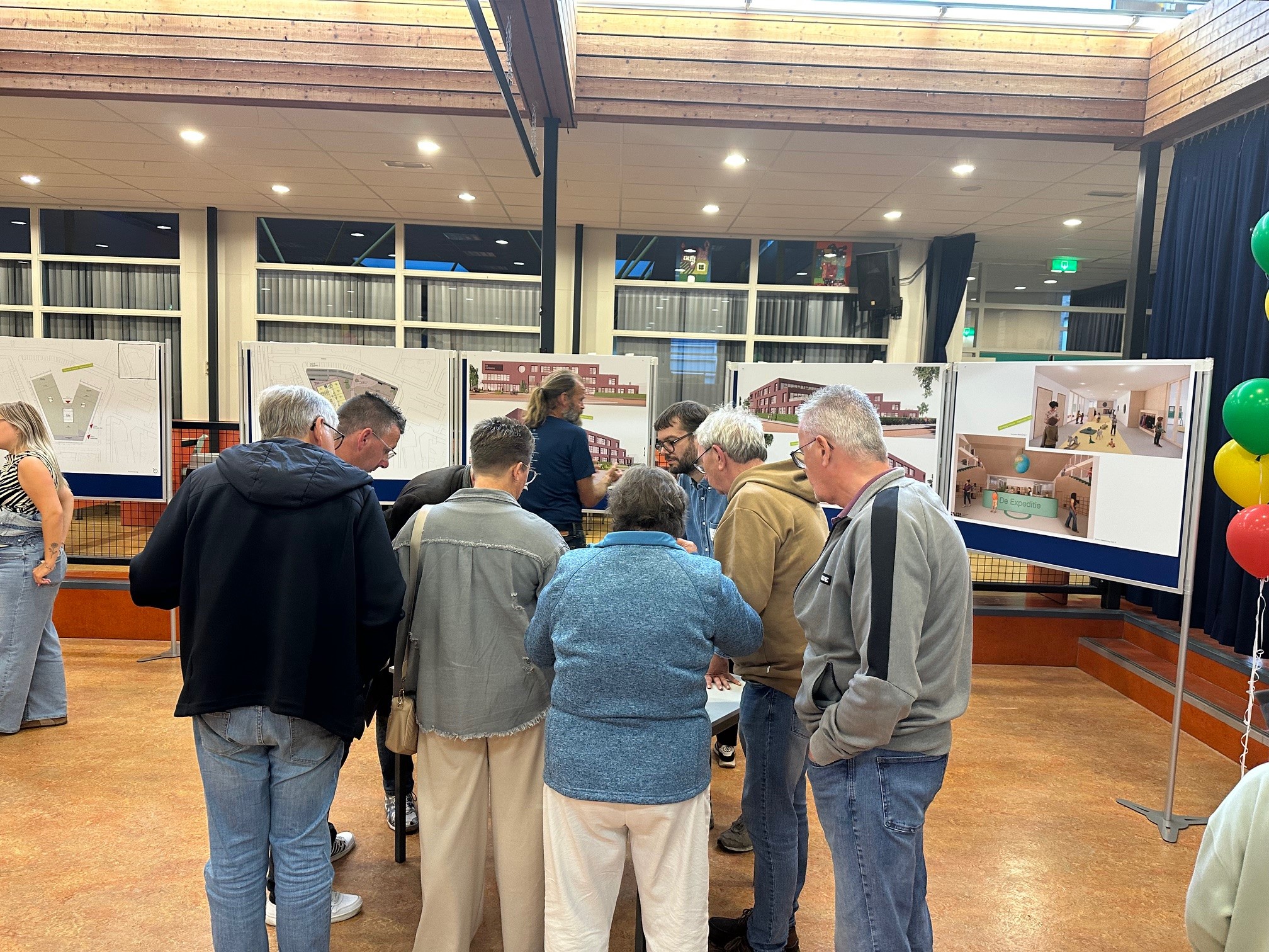 ISW - Een groep mensen staat in een ruimte en bekijkt posters met architectuurtekeningen en interieurrenderings voor het voorlopig ontwerp van Expeditie Poeldijk. Kleurrijke ballonnen rechts dragen bij aan de levendige sfeer van deze publieke presentatie.