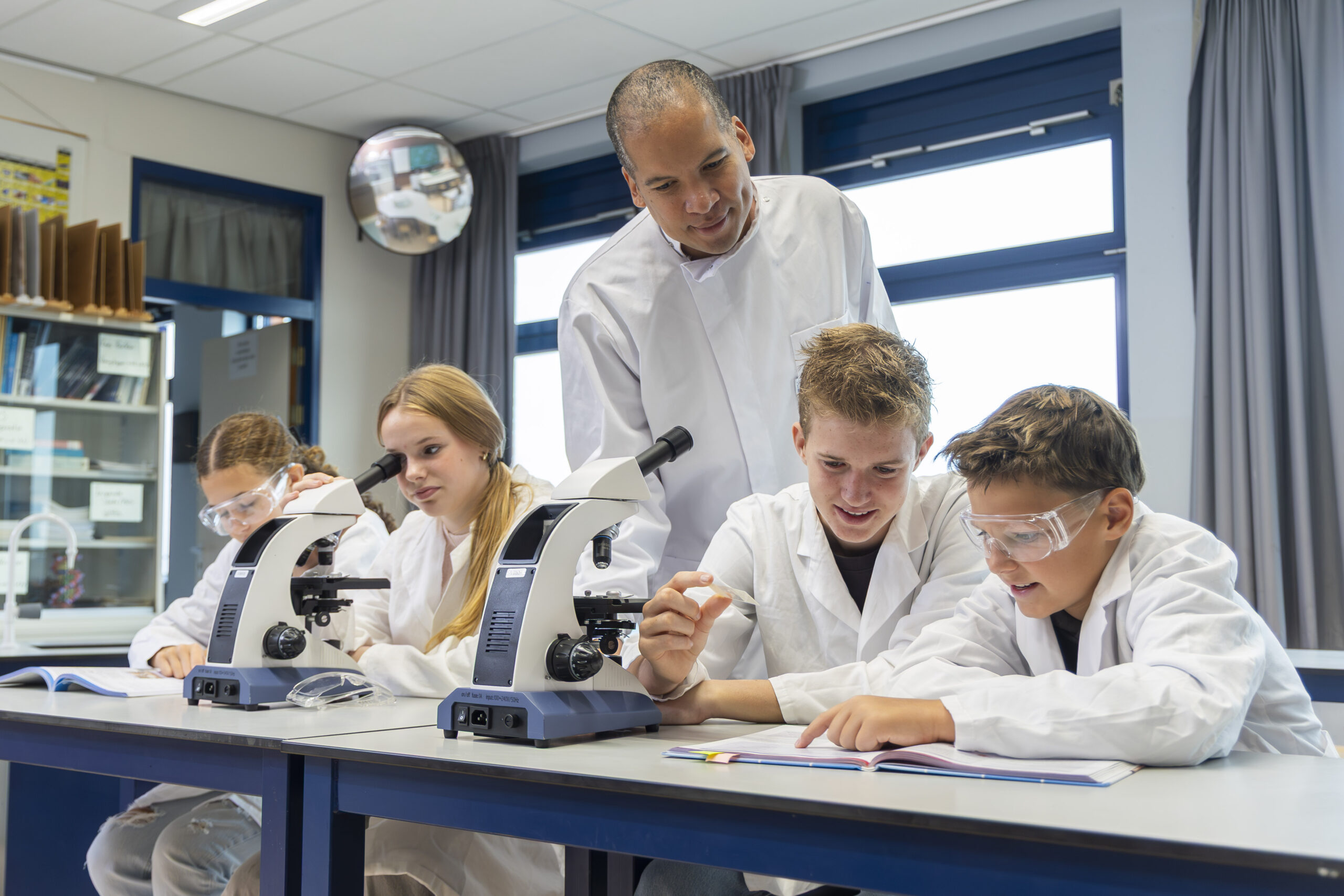 ISW - Een docent in een labjas begeleidt vier studenten, ook in labjas en veiligheidsbril, terwijl ze met microscopen werken en een boek lezen in een helder wetenschapslokaal.