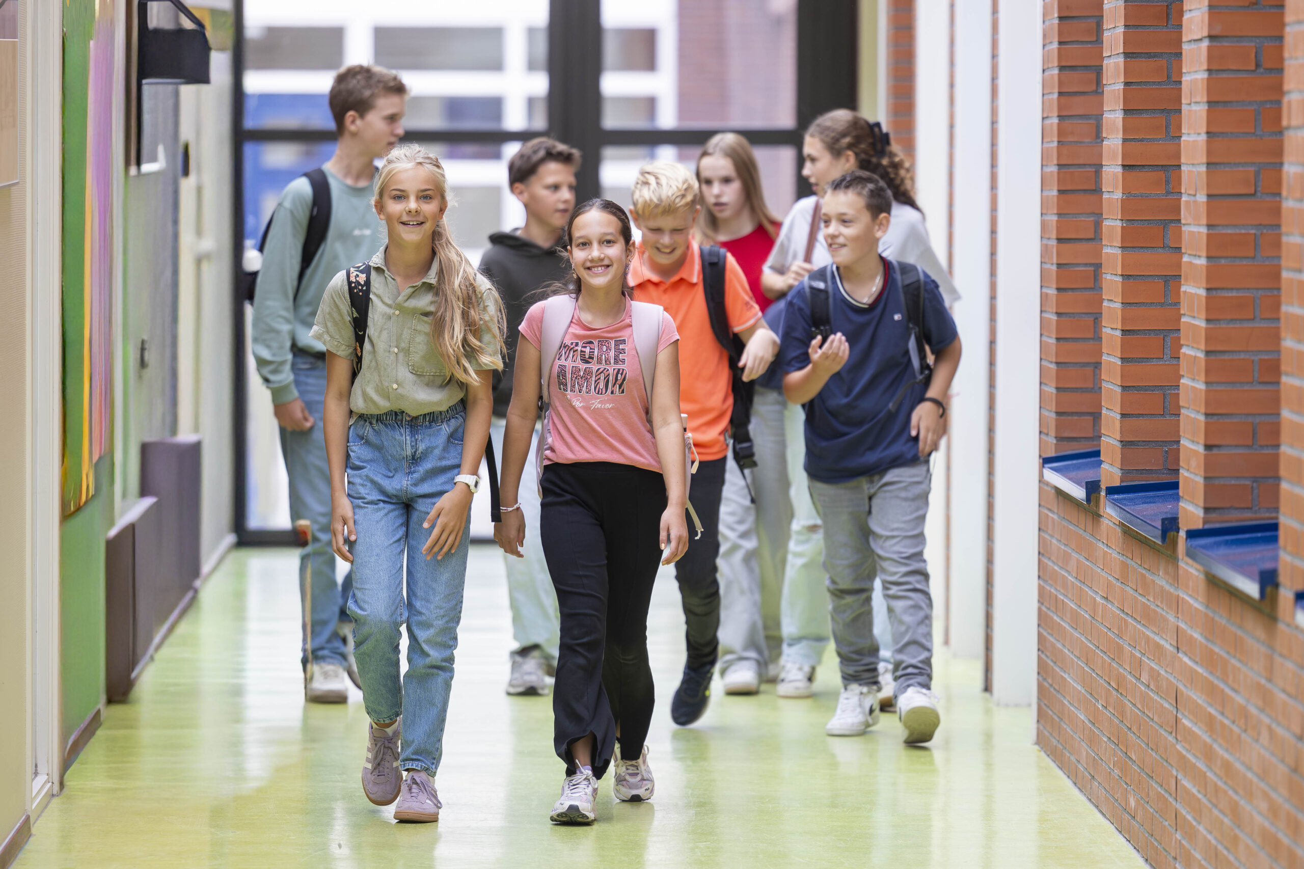 ISW - Een groep lachende leerlingen met rugzakken loopt door een lichte hal van de school, samen kletsend en lachend. De gang heeft bakstenen muren en grote ramen, die een levendige en uitnodigende sfeer creëren.