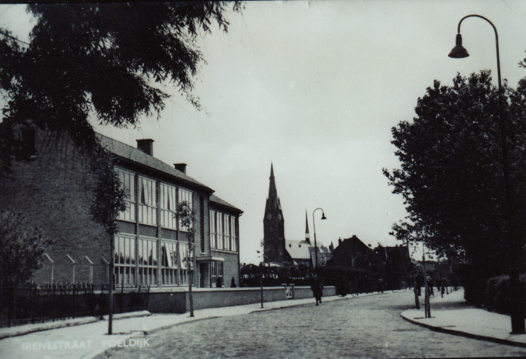 ISW - Een zwart-wit foto van een rustige straat met bomen en gebouwen, waaronder links de ISW Irenestraat. Een kerk met een hoge torenspits staat op de achtergrond; een paar mensen lopen over de stoep tijdens het 70-jarig jubileum van de school.