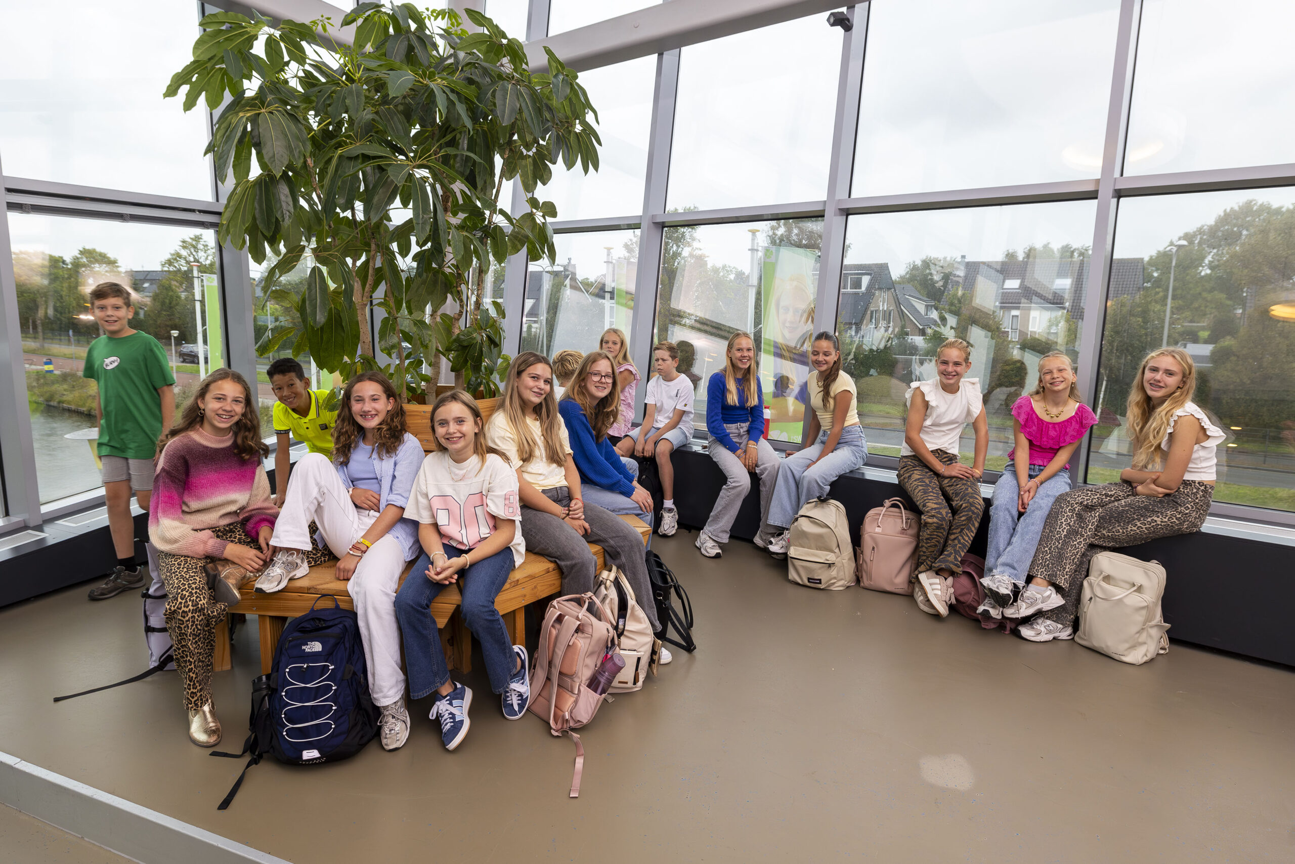 ISW - Een groep tieners zit samen in een lichte, moderne kamer met grote ramen en een hoge plant en lacht naar de camera. Ze hebben rugzakken en vrijetijdskleding.
