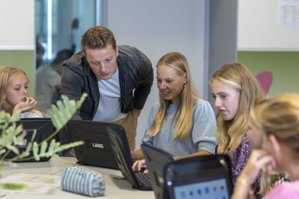 ISW - Een leerkracht leunt voorover om een lachende leerling te helpen die op een laptop werkt, terwijl drie andere leerlingen in de buurt zitten en ook een laptop gebruiken, in een lichte klas.